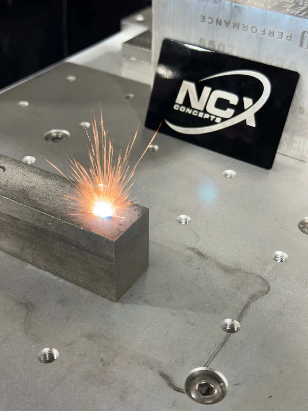 Laser marking a steel block with sparks, beside an NCX Concepts logo plate.
